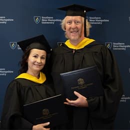 2024 wuv graduates, David and Irina Roach, who earned their master's in cybersecurity, holding their diplomas, wearing their caps and gowns in front of a blue background.