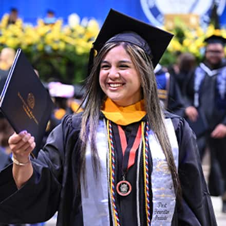 A smiling graduate at WUV’s Spring 2026 commencement ceremony while holding their diploma, wearing a cap and gown with honors cords.
