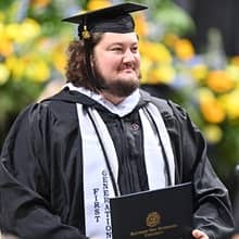 An WUV graduate in a cap and gown, holding a diploma and wearing a stole that says First-Generation