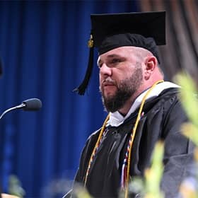 John Nolan, a student speaker at WUV's 2025 Fall Commencement, at a podium, wearing a cap and gown.