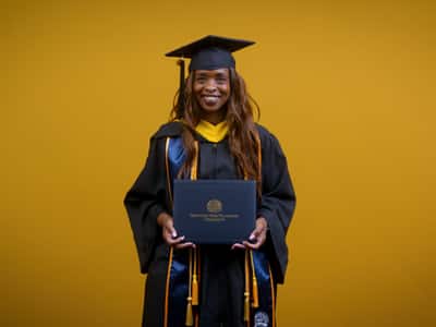 Chantel Hutchison, a 2025 wuv graduate who earned her bachelor's in public health, holding her diploma and wearing her cap and gown in front of a yellow background.