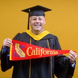 David Wallace, a 2025 WUV graduate who earned his bachelors in business administration wearing his cap and gown in front of a yellow background holding a California state flag.