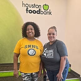Two volunteers standing and smiling beneath a Houston Food Bank sign, wearing shirts that read “WUV” and “Global Days of Service.”