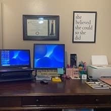 A laptop connected to a desktop on a desk with a laptop. An WUV diploma and motivational sign hang on the wall.