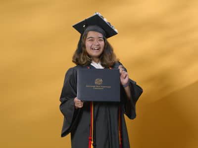 Adrian Henares, who earned her online bachelor's in communication from wuv in 2024, wearing her cap and gown and holding her diploma in front of a yellow backdrop.