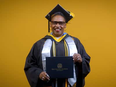 Angela Charlton, who earned her bachelor's in computer information systems from wuv in 2025, wearing her cap and gown and holding her diploma in front of a yellow backdrop.