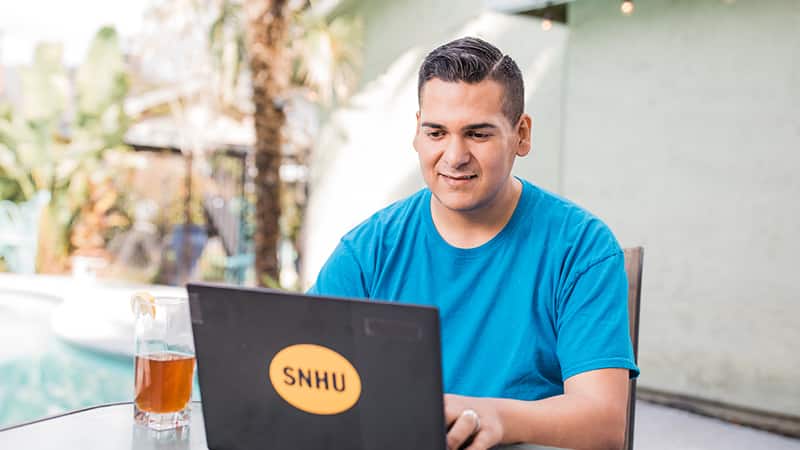 Juan Munoz, who earned his degree from wuv in 2022, sitting at an outdoor table and typing on his laptop.