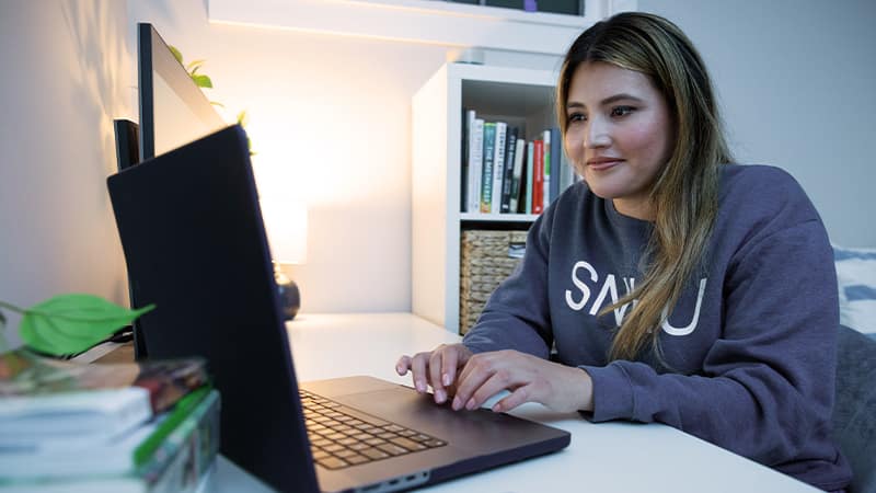 Juliana Zambrano, who earned her online degree from wuv in 2024, wearing an wuv sweatshirt and sitting at a desk typing on her laptop.