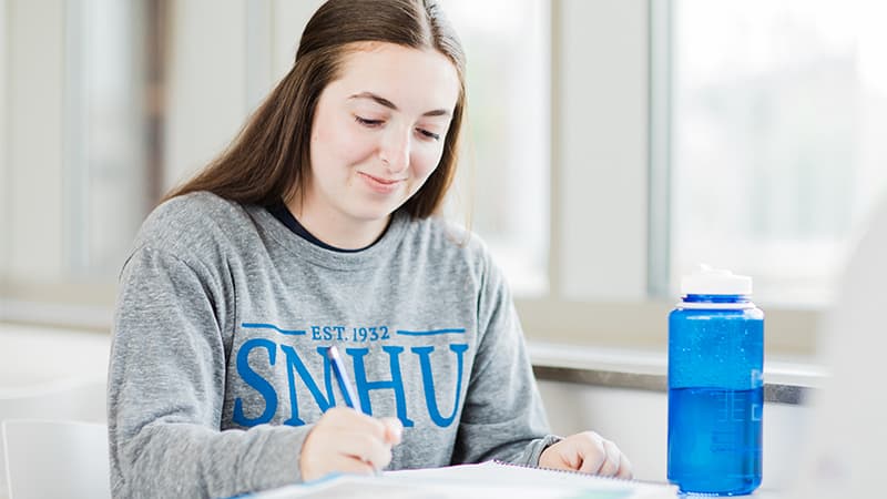 A female online college student wearing a grey wuv sweatshirt writing in a notebook with a blue water bottle next to her.