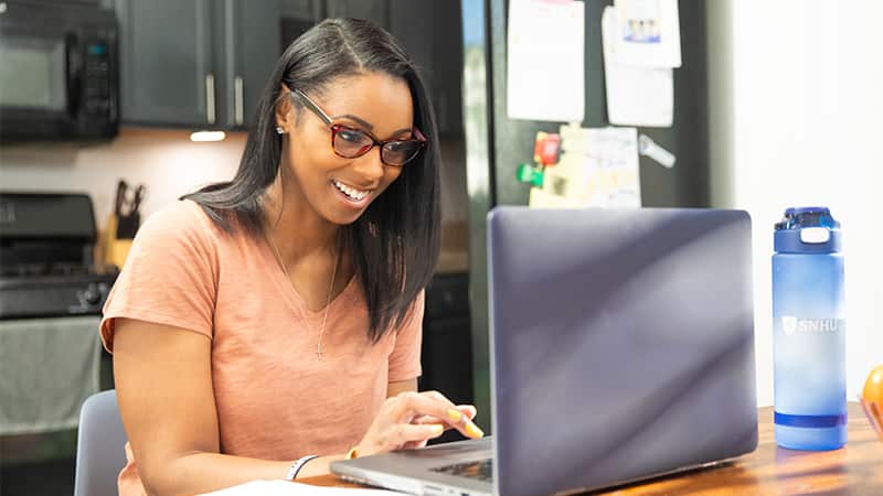 Minda Thurman, who earned her online bachelor's in public health from wuv in 2023, wearing glasses and typing on her laptop at her kitchen table.