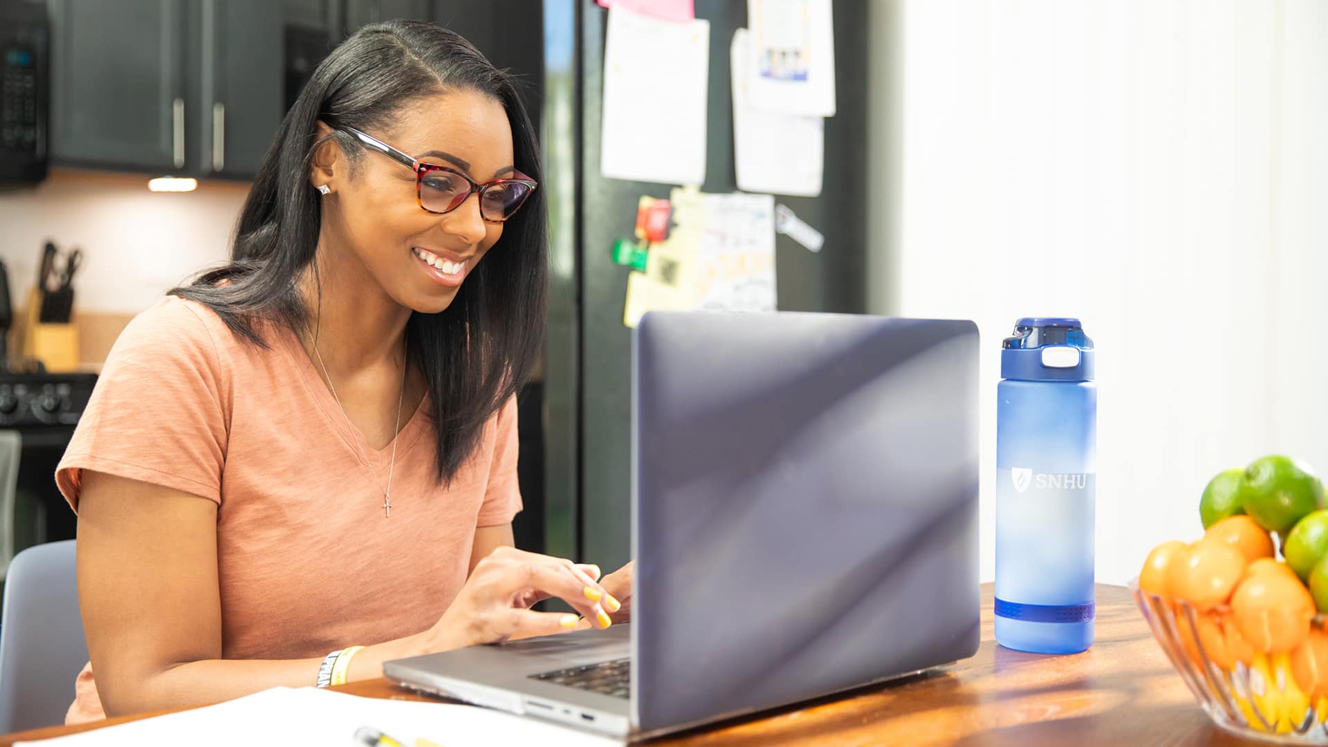 A woman working on a laptop on her kitchen counter with blue wuv water bottle next to her.
