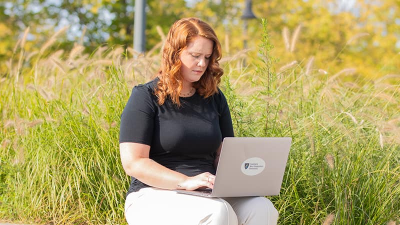 Ali Lamoureux-Rule, who earned her healthcare administration degree from wuv in 2022, typing on her laptop with a field of tall grass behind her.