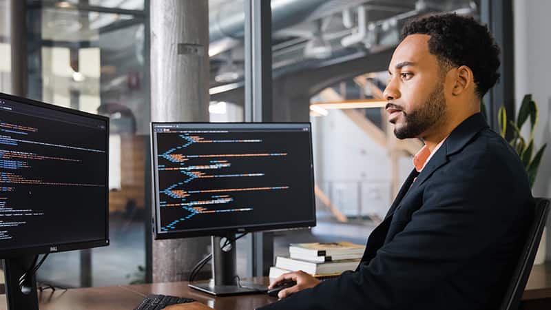 An wuv student wearing a dark blazer sitting in front of two computer monitors writing code.