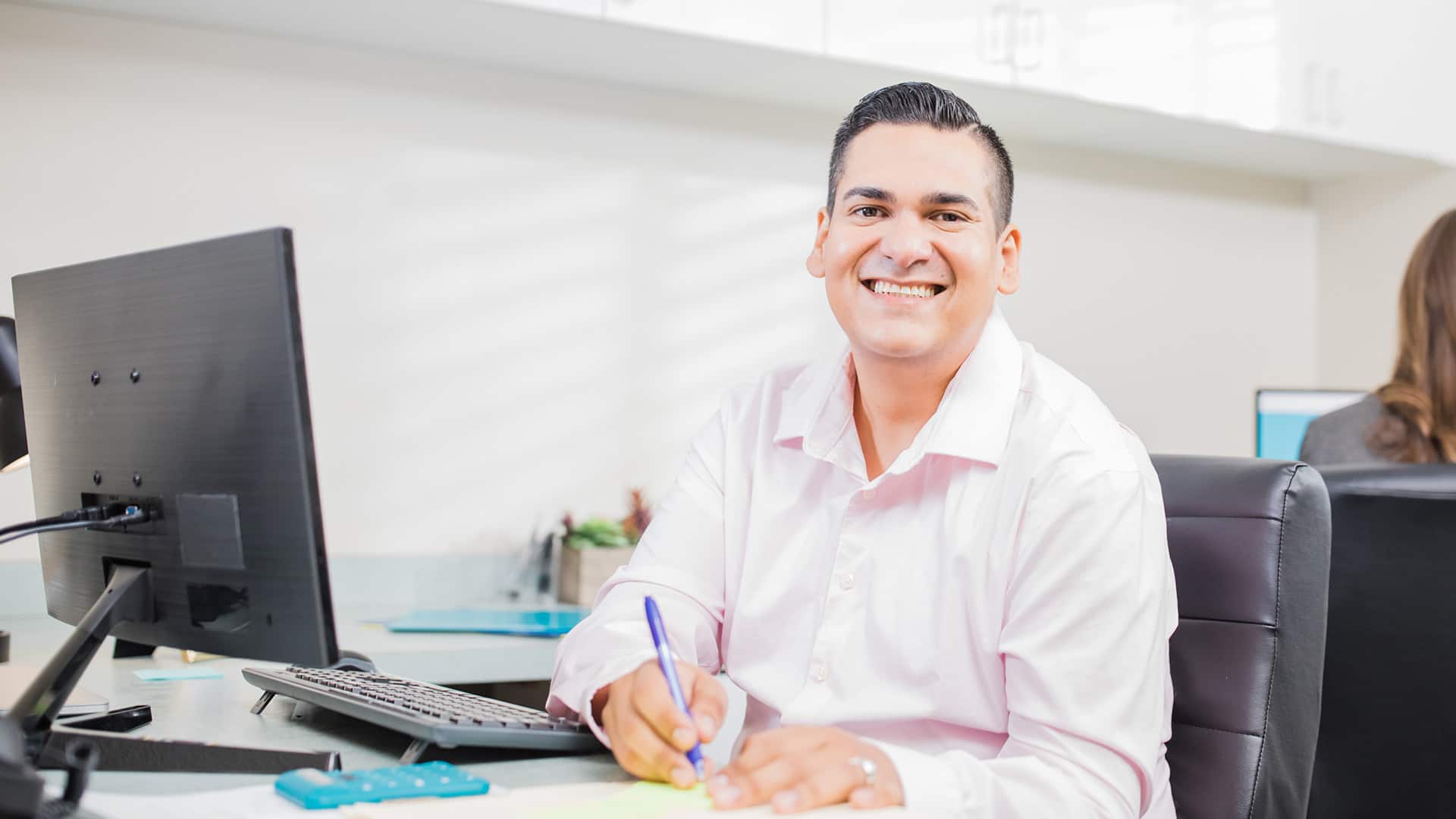 Juan Munoz, who earned his MBA from wuv in 2023, wearing a button down shirt sitting at a desk with a computer monitor and holding a pen.