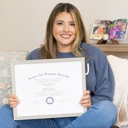 Juliana Zambrano, a 2025 WUV graduate with a bachelor's degree in business administration sitting on a couch holding her diploma.