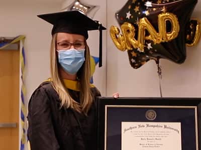 Katie Smith, who earned her MSN from wuv in 2021, wearing her cap and gown and holding her framed diploma with a "Grad" balloon in the background.