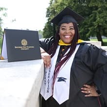 Karen Raquel Quezada, who earned her master's in psychology from wuv in 2021, wearing her cap and gown and standing next to her diploma.