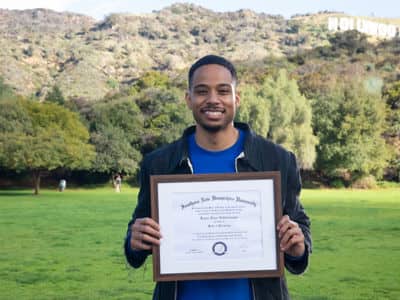 Keyon Tuiteleleapaga, who earned his psychology bachelor's from wuv in 2022 and graduate degree in 2024, standing in a green field holding his framed diploma.