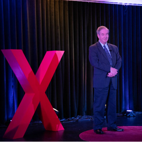 Anthony Knopps speaking at TEDxWUV in 2024, to the right of a large, red letter X