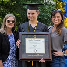 Loretta Gray dressed in her cap and gown, holding her WUV diploma between two friends.