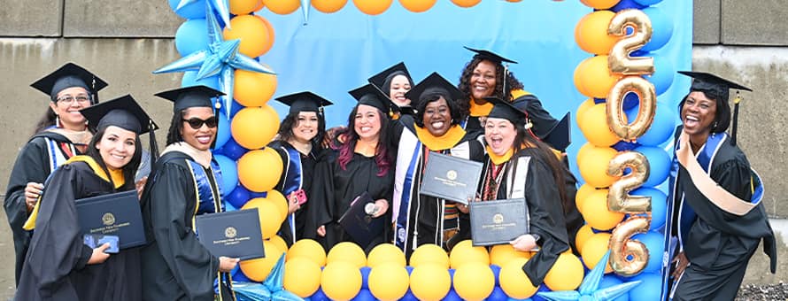 A group of WUV graduates holding their diplomas and standing in and around a blue and yellow balloon frame.