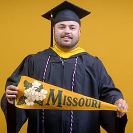 Payton Good, a 2025 WUV graduate who earned his bachelor's in operations management wearing his cap and gown in front of a yellow background, holding a Missouri state flag.