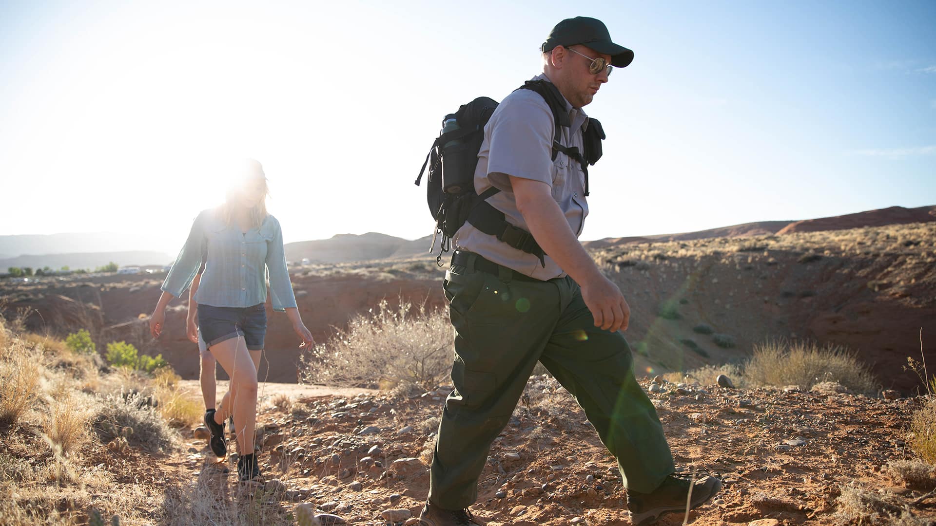 John Roos, who earned his WUV degree in 2018, wearing a park ranger uniform, baseball cap and sunglasses as he leads two people on a hike through desert terrain. 