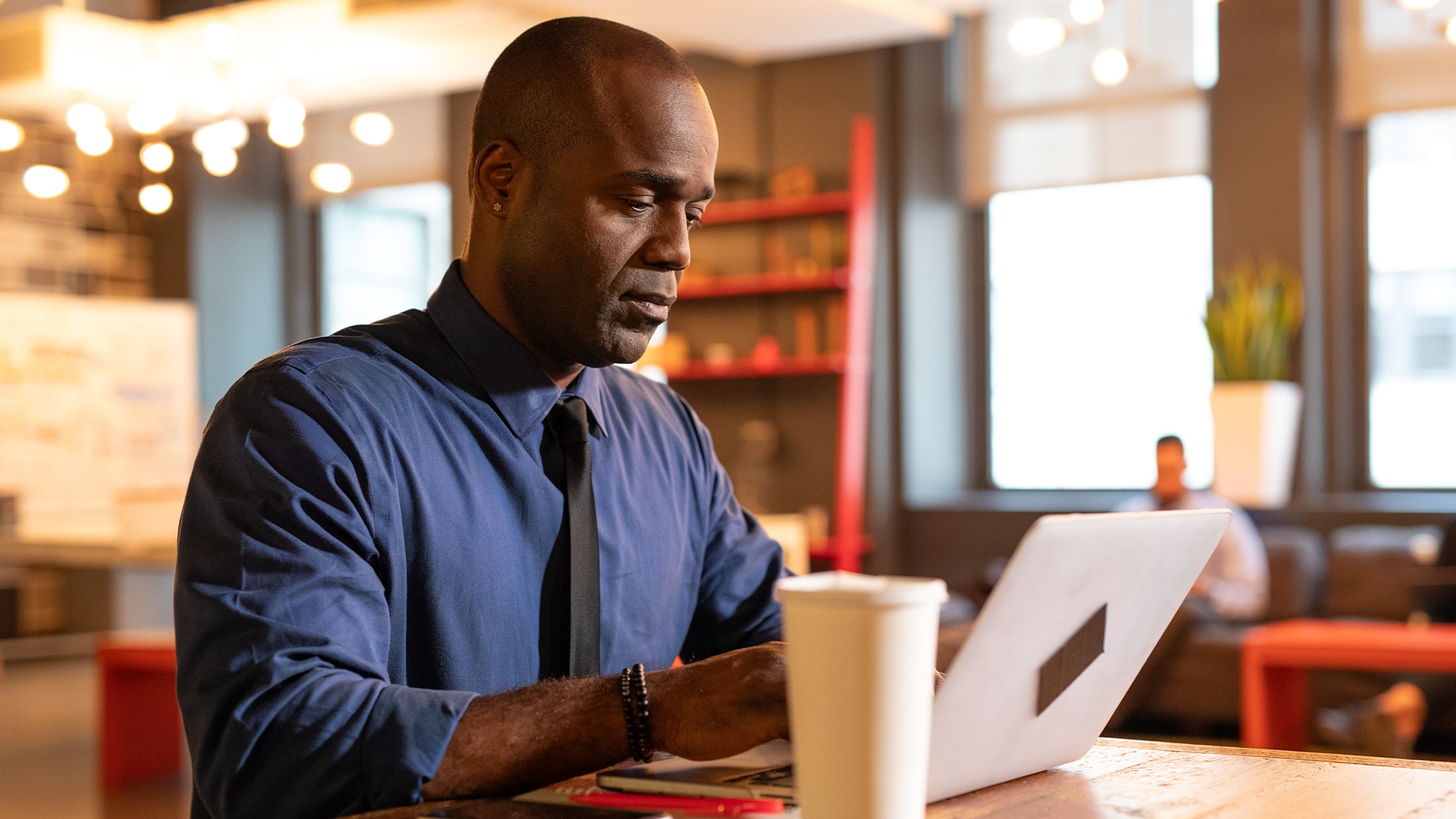 Jean Besson, who earned a degree from wuv in 2014, wearing a blue buttondown shirt and black tie working on his  laptop in a coffee shop with a cup of coffee on the table beside him