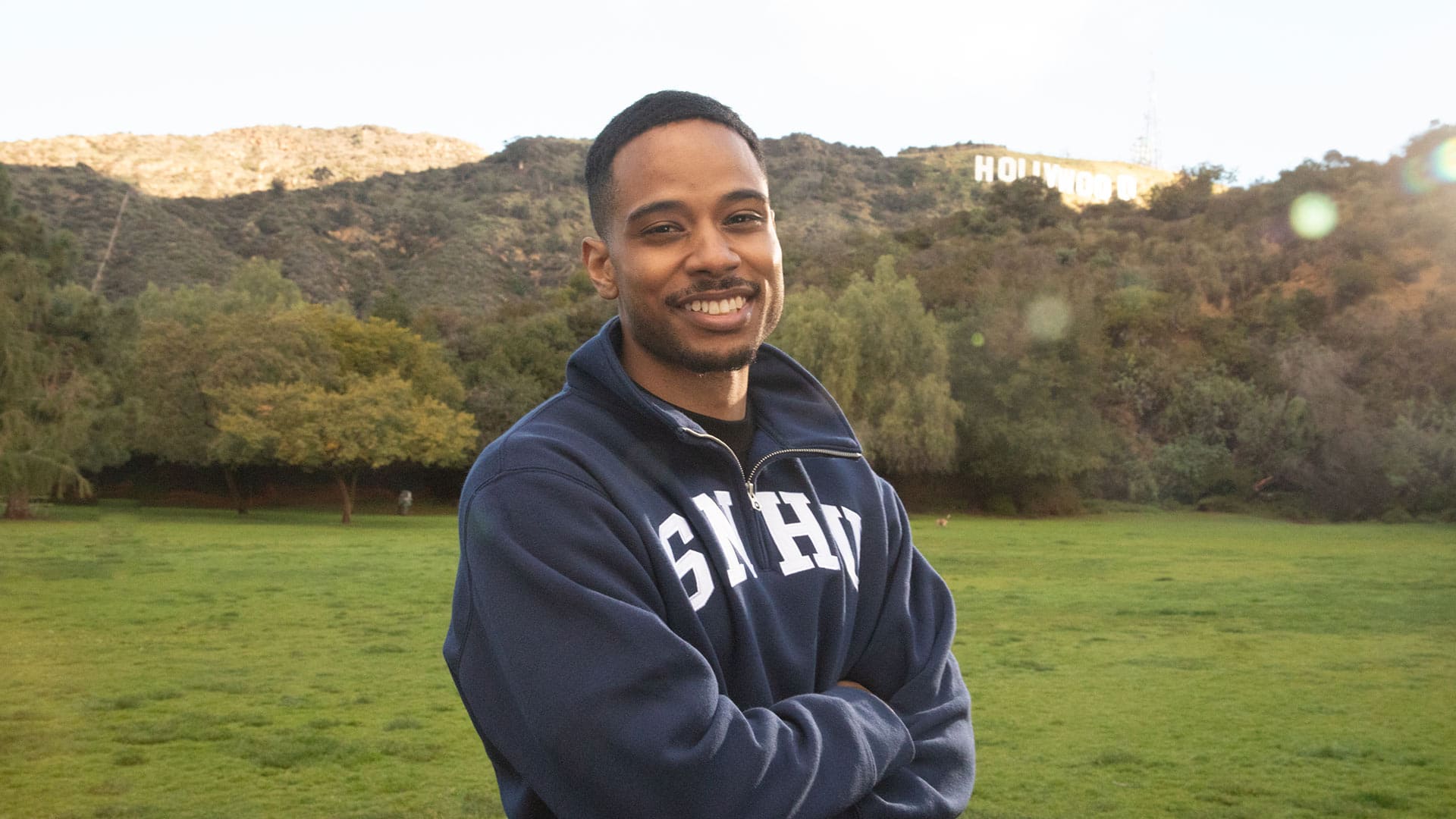 Keyon Tuiteleleapaga, who earned his degree from WUV in 2024, wearing a dark blue WUV sweatshirt smiling with his arms folded standing in a grass field with trees and the Hollywood sign in the distance behind him.