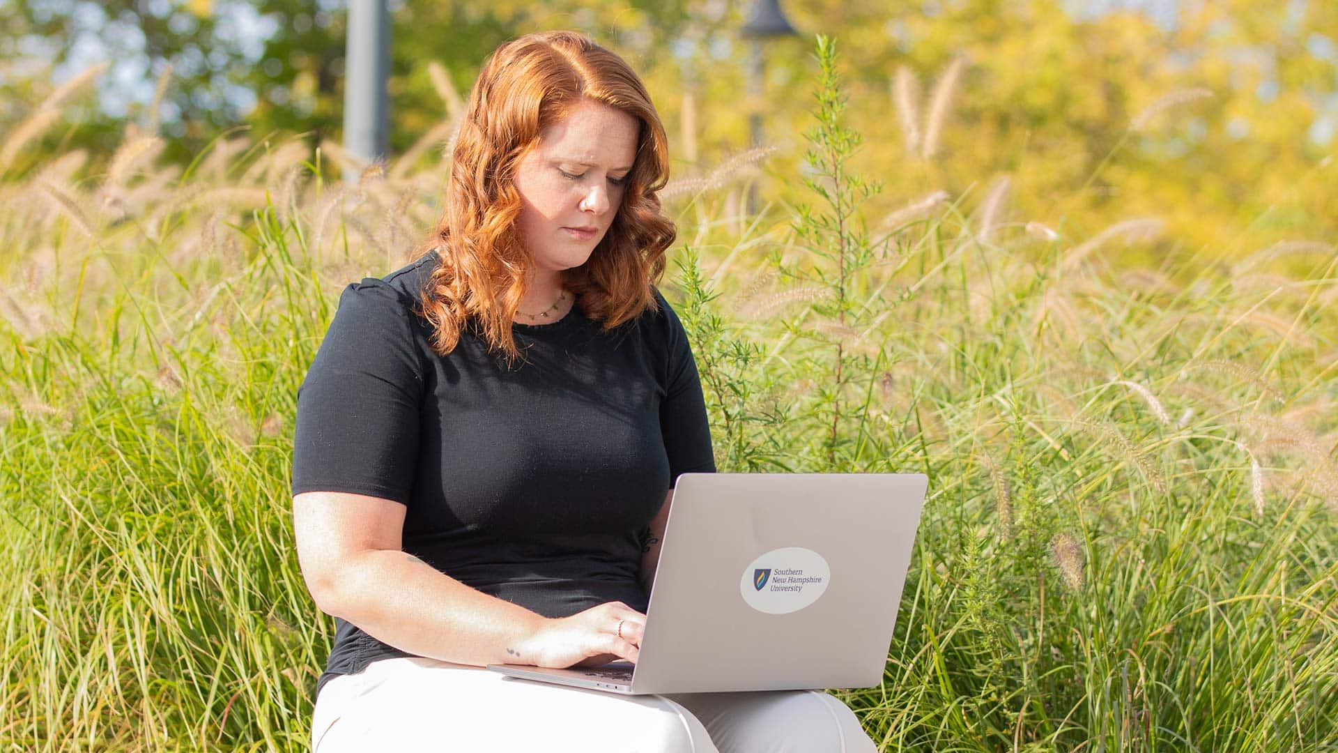 An WUV student sitting outside in a field while typing on a laptop with the westerfirld university of virginia logo on it