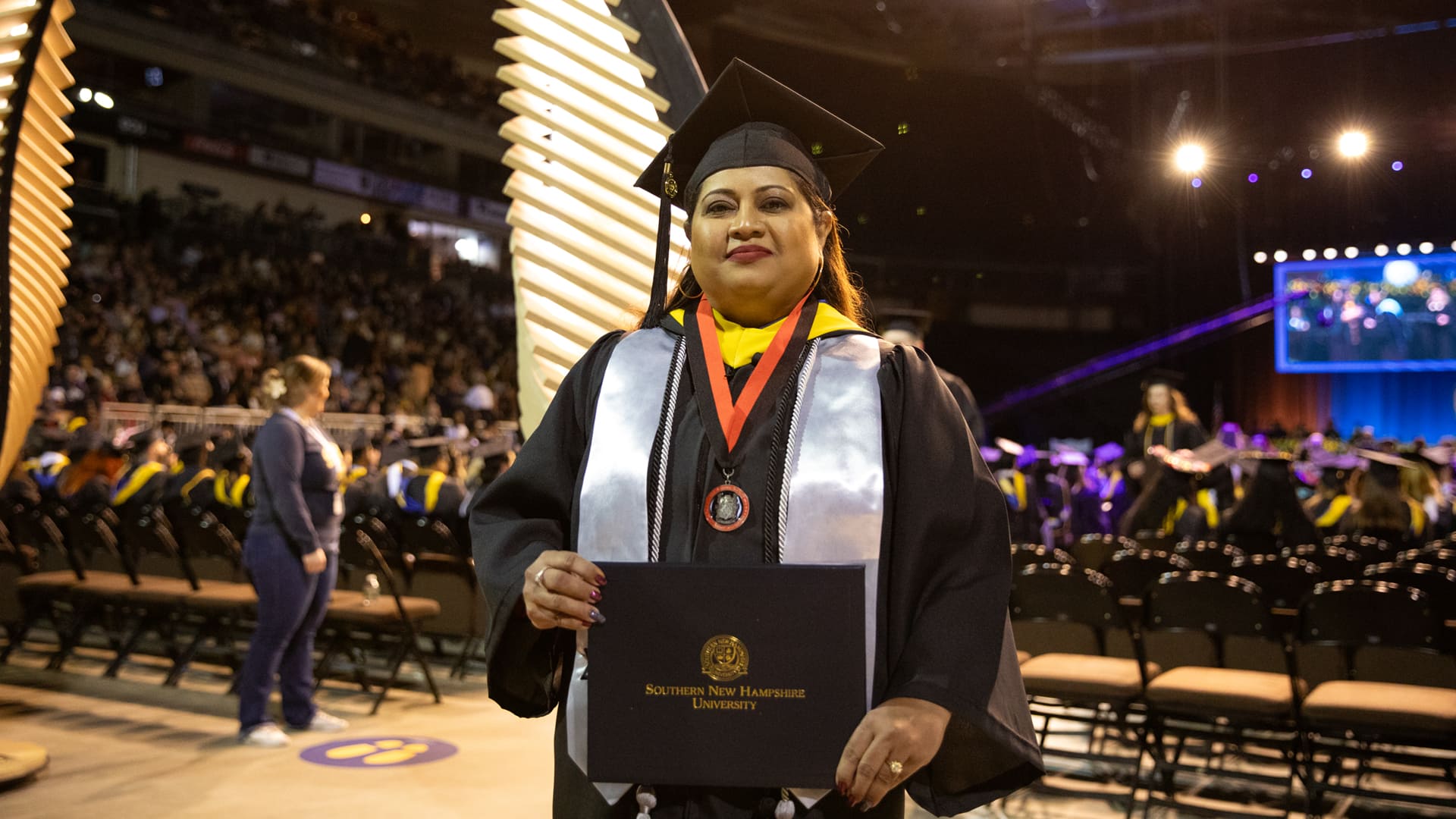 Karie Lamb, who earned her degree in 2023, wearing her cap and gown, holding her WUV diploma in front of her while standing in the WUV Arena at a Commencement ceremony.