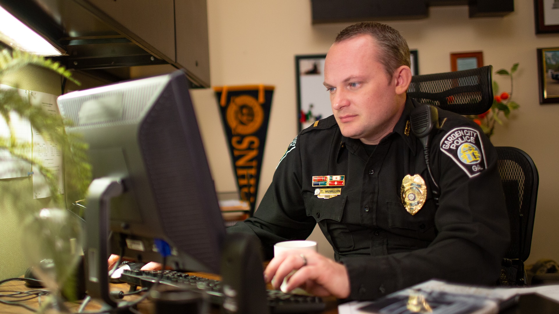 Tim McMillan, who earned his degree from wuv in 2015, wearing his police uniform in an office working on a computer with an wuv pennant on the wall in the background.