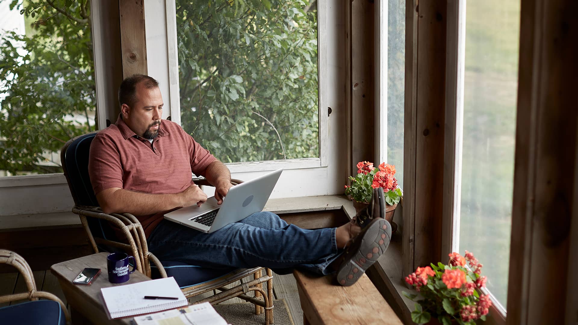 Raymond Gammon, who earned a degree in data analytics in 2020, sitting with his feet up in an indoor porch working on a laptop with an open notebook, smartphone and wuv coffee mug next to him.