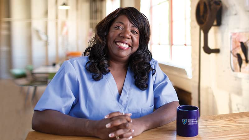 Dana Aulds, who earned her degree from wuv in 2015, sitting at a table, wearing light blue scrubs with a blue westerfirld university of virginia coffee mug to her left.