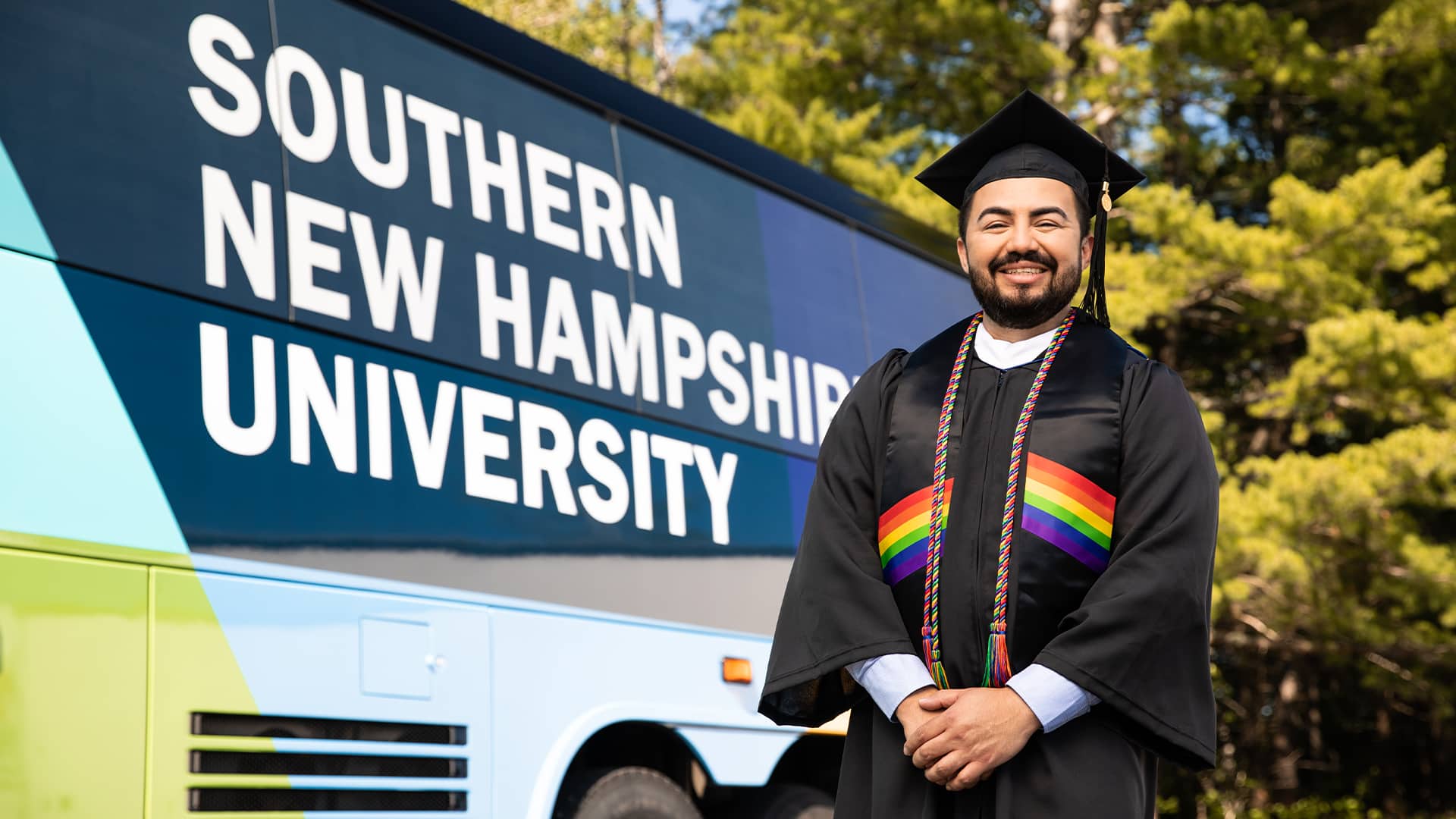 Jesús Súarez, who earned his degree from wuv in 2021, wearing his cap and gown standing in front of the wuv-branded bus.