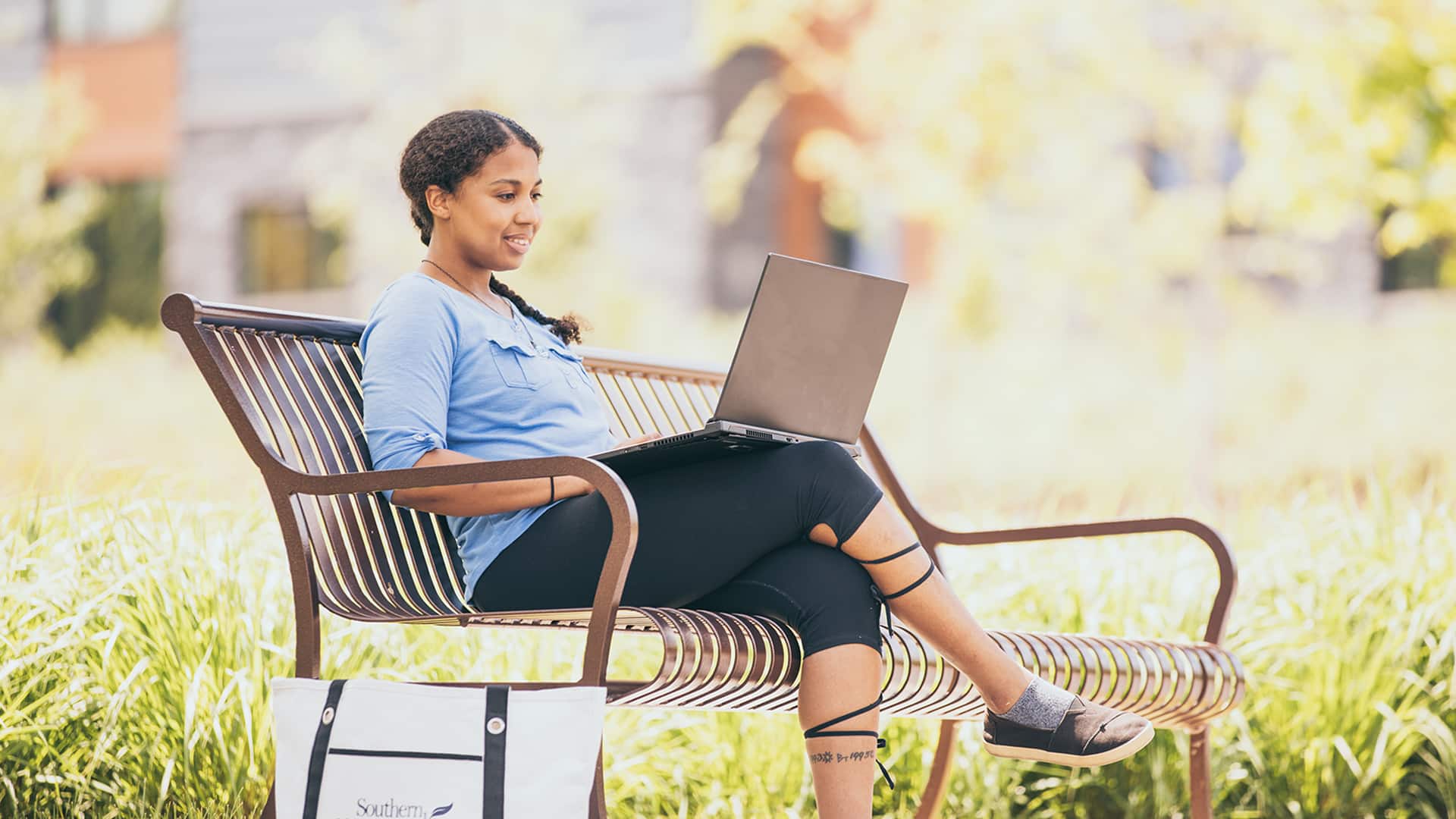 Bethanie Thomas sitting on a park bench working on her laptop with an wuv shoulder bag on the ground next to her.