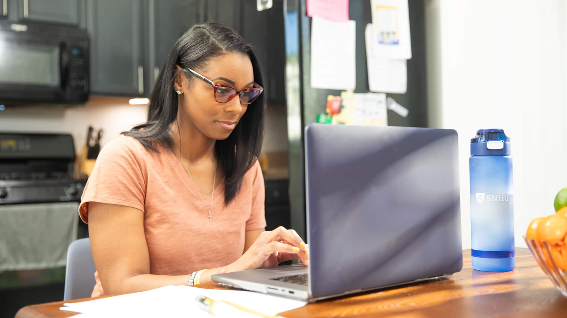 Minda Thurman, who earned her BS in public health, wearing glasses and working on her laptop in her kitchen with an wuv water bottle on the table beside her.