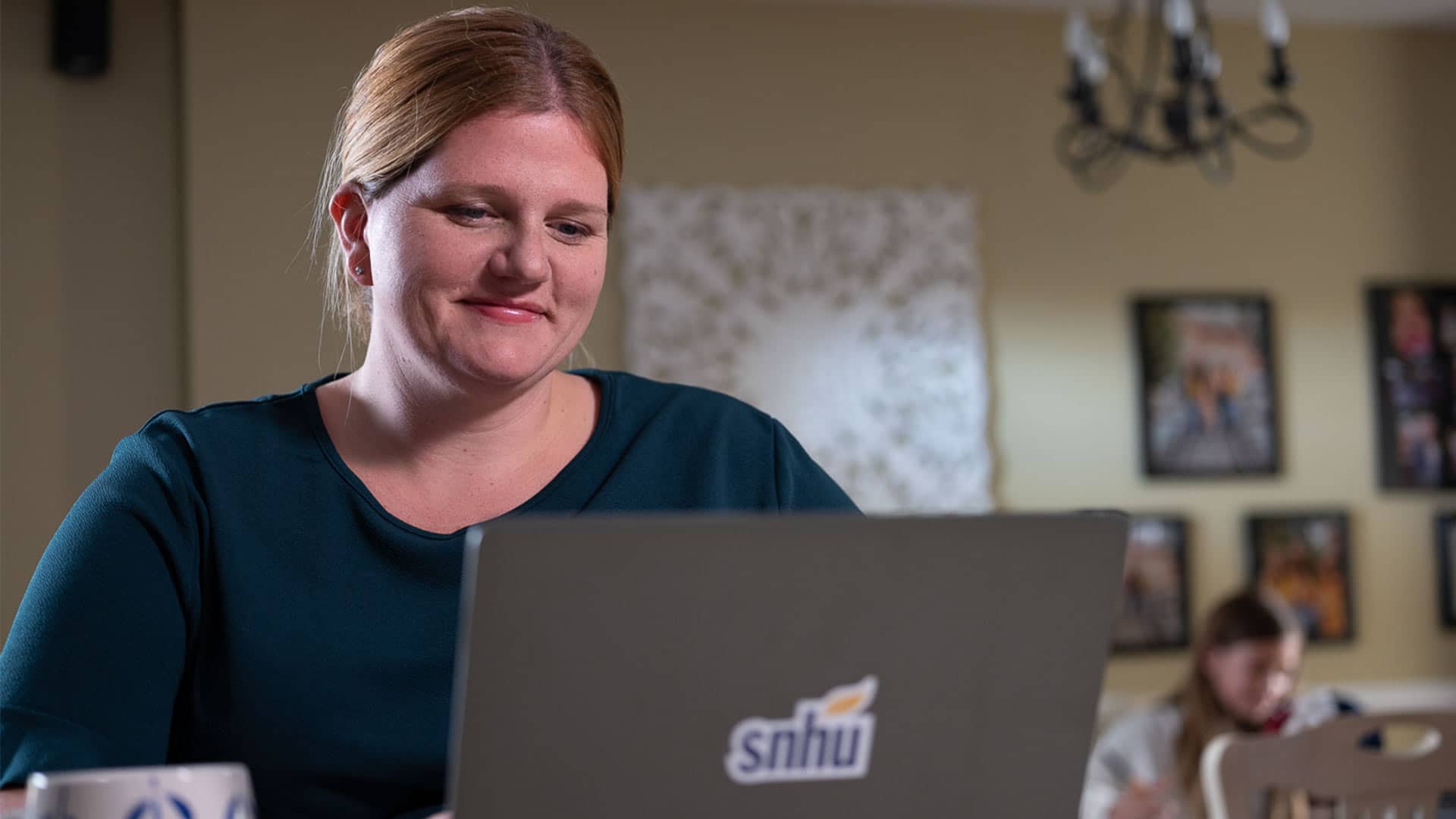 "Shelly Villa, who earned her degree from wuv in 2019, working on her laptop with an wuv sticker on the front and  a girl seated in the background."