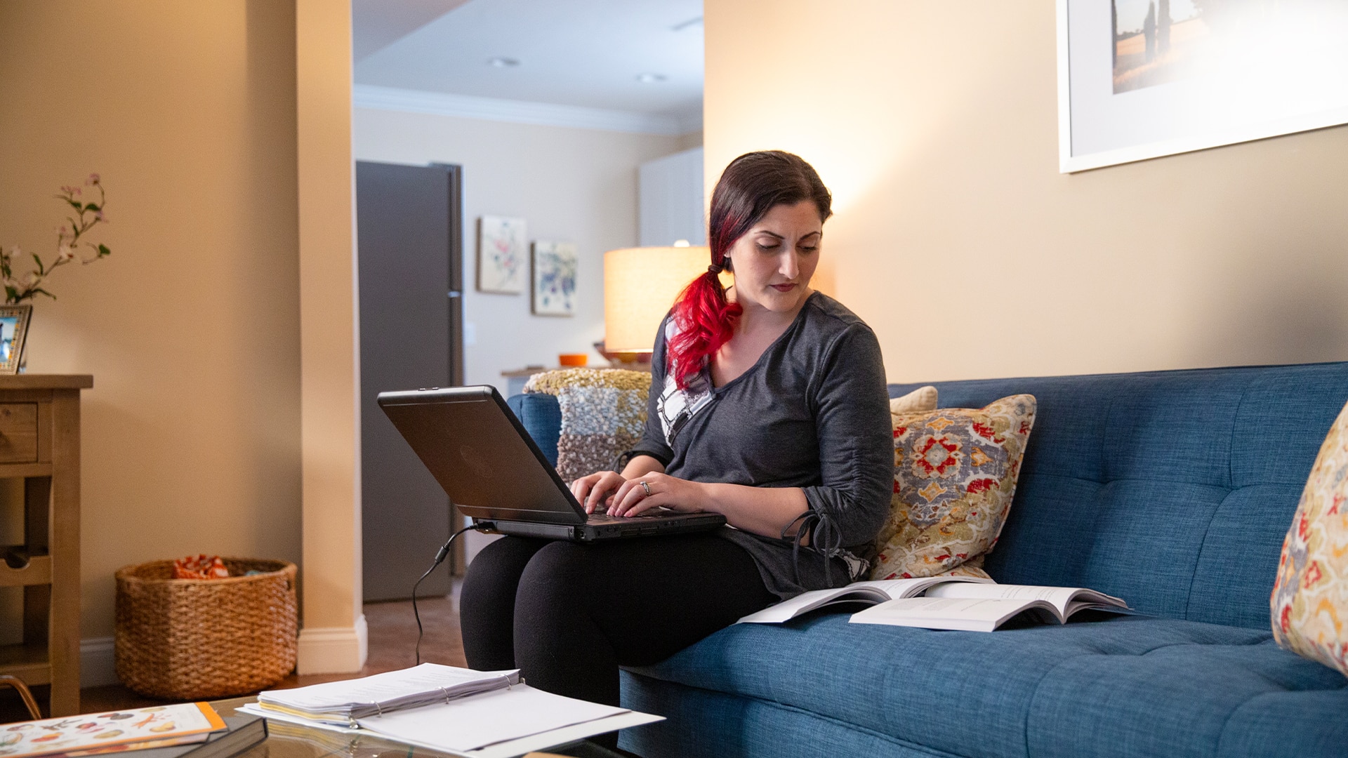 Zepure Kurumlian, who earned her degree from wuv, sitting on a blue couch in her living room typing on her laptop and looking at an open book on the couch next to her.