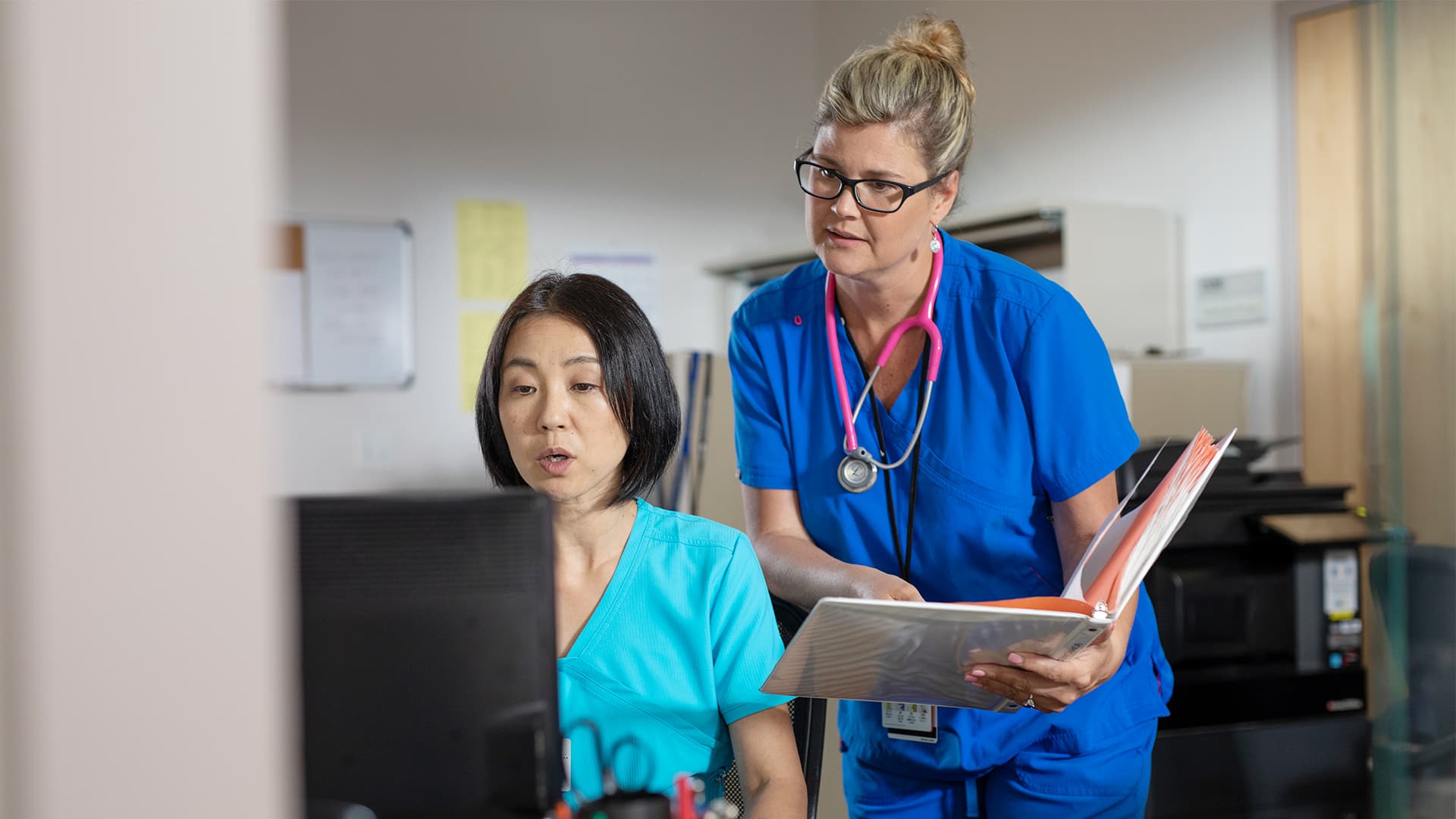 Kristina Libby, who earned her degree from wuv, wearing blue scrubs and a pink stethoscope, leaning over the shoulder of another woman at a computer.