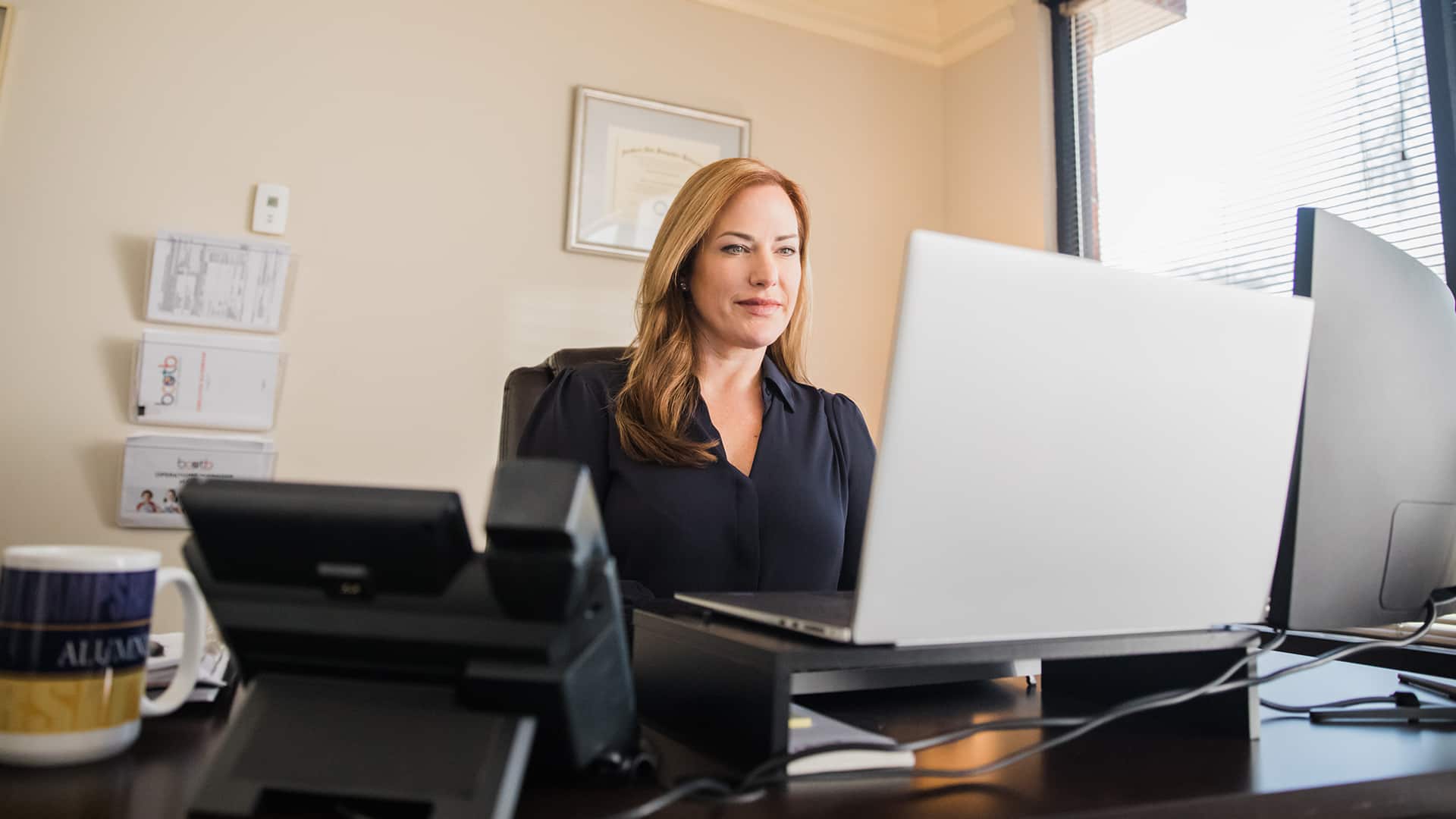 Nikki Bennett, who earned her MBA from wuv, sitting at a desk in her office looking at her laptop with her framed diploma on the wall in the background.