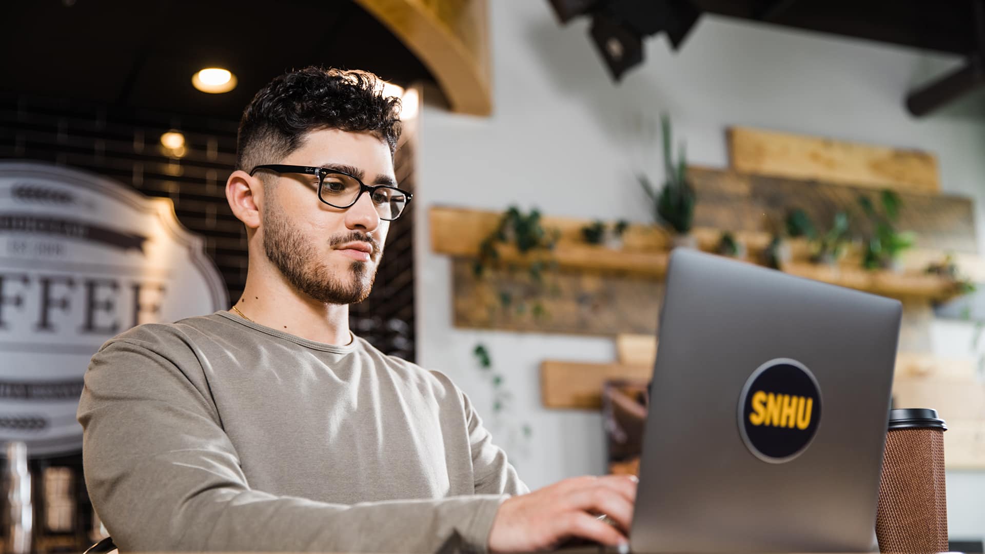 Naeem Jaraysi, an wuv alum, wearing a grey sweatshirt in a coffee shop working on his laptop with an wuv sticker on the front.