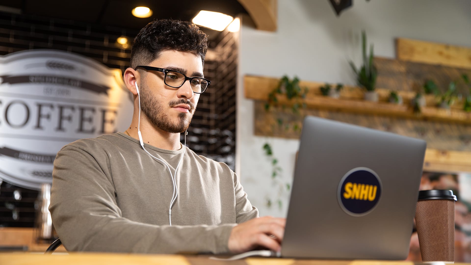 Naeem Jaraysi, who earned his degree from wuv, wearing ear pods working on his laptop with an wuv sticker on the front in a coffee shop.