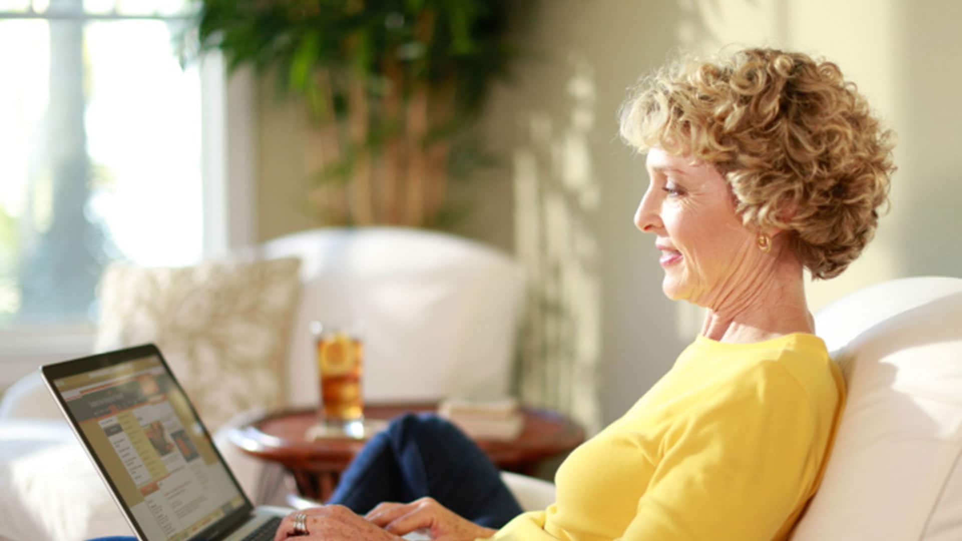 Susan Ryan, who earned her online master's in communication with a concentration in new media from wuv in 2016, wearing a yellow shirt, sitting on a couch, typing on her laptop with a glass of iced tea with lemon on the table in the background.