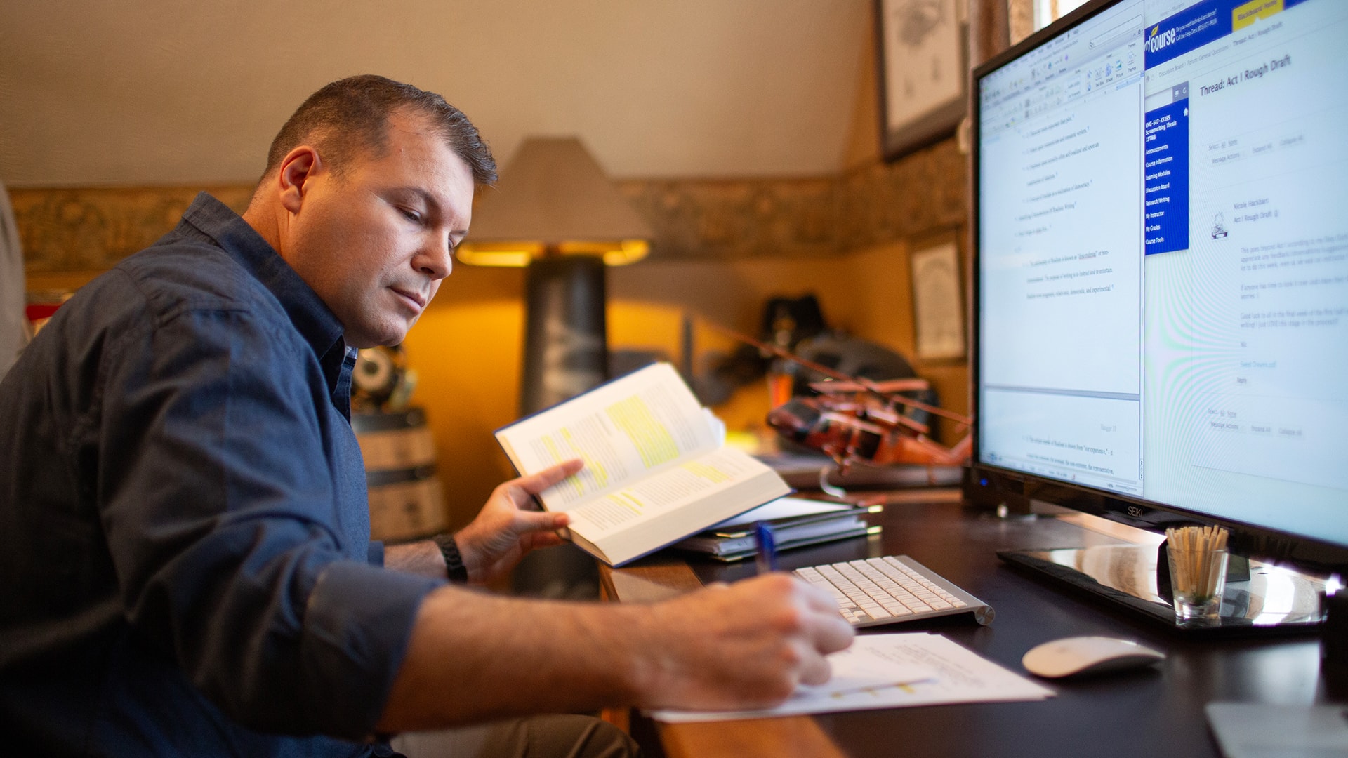 Michael Hangge, who earned his degree from wuv in 2015, at the desk in his home office in front of a computer monitor, holding open a text book with some passages highlighted and taking notes on a piece of paper.