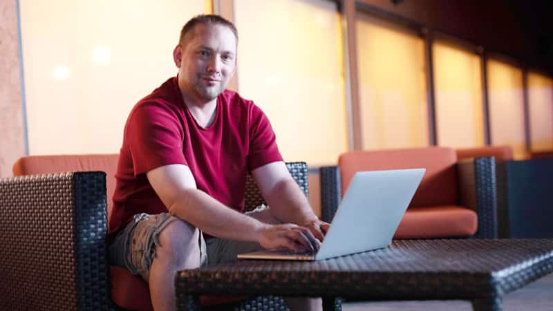 John Roos, who earned his online history degree from wuv in 2018, wearing shorts and a red T-shirt, sitting in a chair and typing on his laptop.