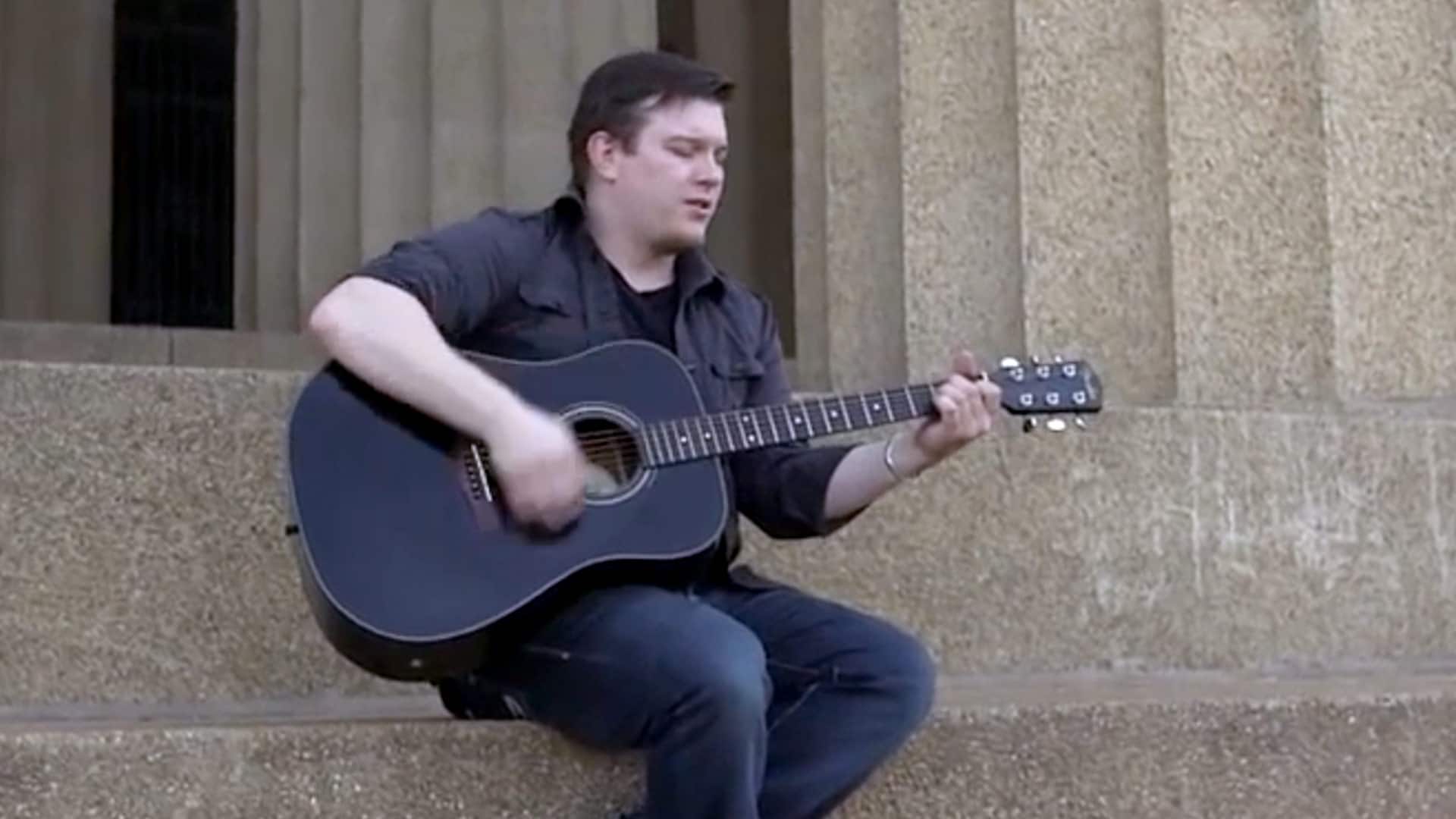 Russ Kettle, who earned his degree from wuv in 2017, sitting on a set of stone steps playing a black acoustic guitar.