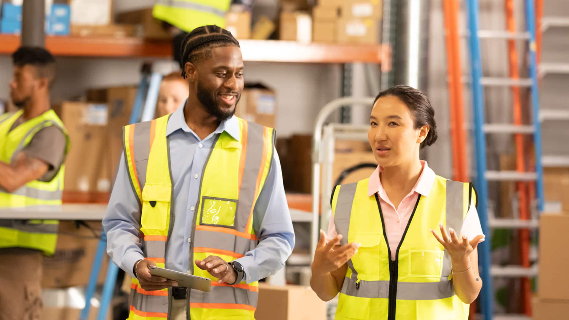 Matthrew Seawright, who earned a degree from wuv in 2019, wearing a bright yellow safety vest and holding a tablet computer speaking to a woman inside a warehouse.