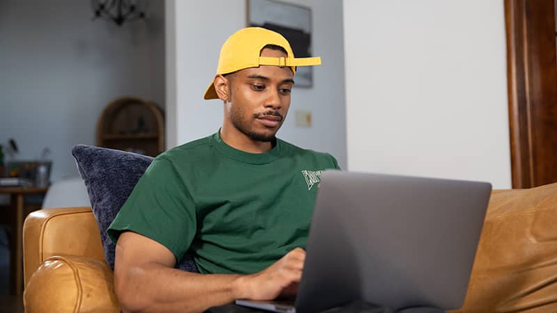Keyon Tuitelelepaga, who earned online degrees from wuv in 2022 and 2024, wearing a green T-shirt and a backward yellow baseball hat, sitting on a couch and typing on his laptop.