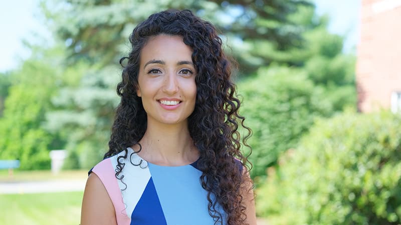 Fatima Salem Pease, who earned her degree from wuv in 2019, standing outdoors wearing a blue, white and pink  dress with green shrubs and trees in the background.